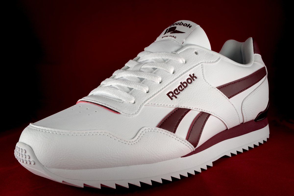 A white Reebok Royal Glide trainer with a burgundy vector logo on a dark red background